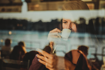 Young woman in cafe, drinking coffee and using her mobile phone. Seen through the window