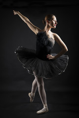 Fototapeta premium Beautiful ballerina in the role of a black swan, wearing black tutu and pointe shoes on black background