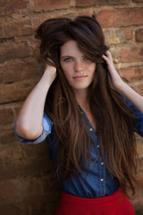 Youngseductive, girl with long brown hair and a birthmark on face in front of brick wall wearing jeans blue shirt 