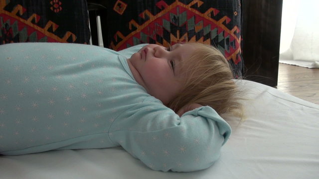 blonde caucasian baby face two years old age with light blue cyan shirt sleepy and closing eyes to sleep on white sheet stretching her arms
