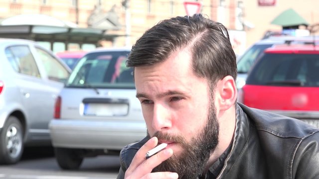 young handsome hipster man smokes a cigarette on the street - parked cars in the background