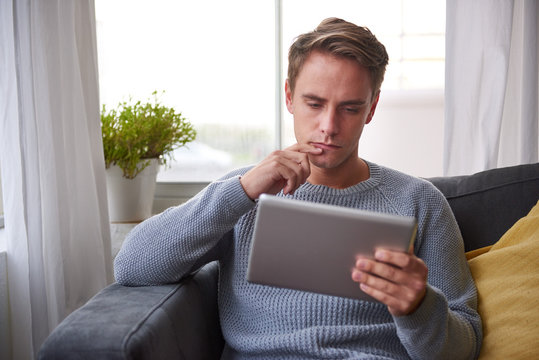 Guy on his couch looking pensive while reading a digital tablet