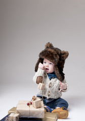 Funny baby in a winter fur hat with gift boxes over gray backgro