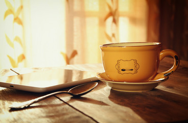 Cup of tea with tablet device next to window on wooden table
