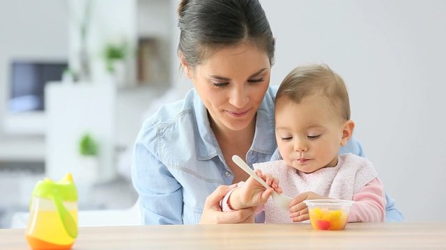 Young mother helping baby girl with eating by herself