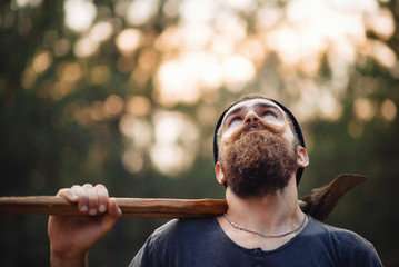 Brutal brunette bearded man in warm hat with a hatchet in the woods on a background of trees