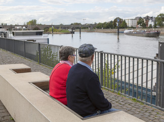 An elderly caucasian couple is sitting on a bench by the river and looking ships passing by.  The woman has a red shirt and the man has a cap on his head.