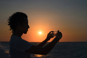 Silhouete of young man taking photo of sunset at seaside with his smartphone