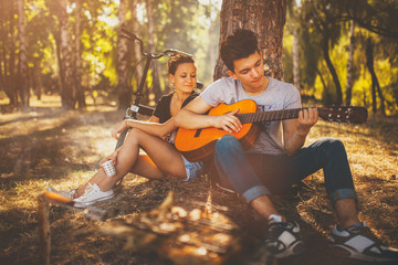 Teen boy sitting with his girlfriend by a tree, having a picnic and playing guitar on a sunny autumn day in forest. Teenagers loving couple relaxing by bonfire outdoors - Powered by Adobe