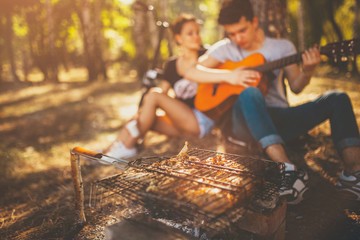 Fototapeta na wymiar Picnic. Grilling meat and playing guitar. Defocused background. Teenagers loving boy and girl sitting together by a tree on weekend sunny day.