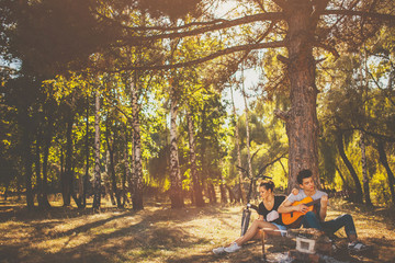Young loving couple having a picnic in forest on a sunny autumn day. Teenage boy and girl playing guitar outdoors by a tree.