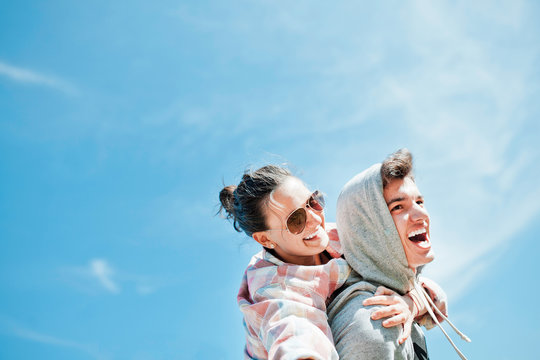 Happy Loving Couple. Happy Young Man Piggybacking His Girlfriend. Cheerful Laughing Hipsters On Blue Sky Background With Copy Space