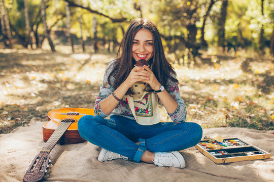 Adorable teenager girl wearing casual clothes having a picnic on autumn day in forest. Cute and beautiful hipster young woman smiling and holding a cupcake, sitting on blanket by guitar.