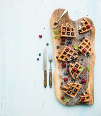 Soft Belgian waffles with berries, honey and mint on rustic wooden serving board over light blue background, top view