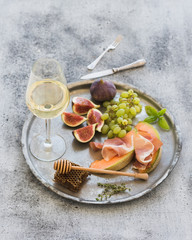 Wine appetizer set. Glass of white wine, honeycomb with drizzlier, figs, grapes, melon and prosciutto on silver tray over rustic grunge surface