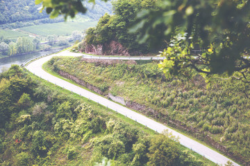 A dangerous and curvy road at the mountains. A narrow road circles around the high hill and below is a river far away. Image has a vintage effect applied.