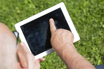 An elderly man is browsing the internet with a tablet outdoor with a green grass on the background.