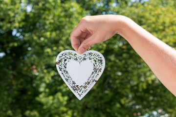 Obraz premium Hand holds hanging heart made of white metal in a park with tree