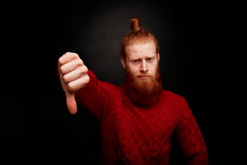 Portrait of bearded man in a red knitted sweater looking at camera and showing his thumbs down while standing against black background.