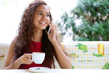 Young smiling woman in red dress speaking by cellphone and drinking coffee at restaurant's terrace