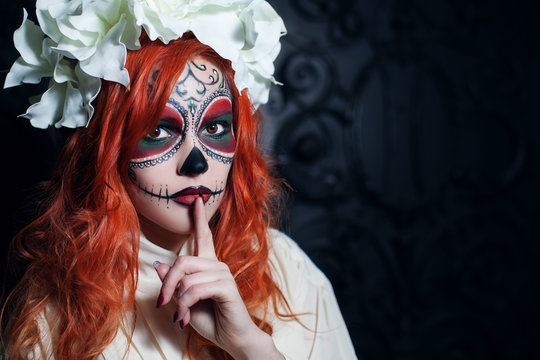 Beautiful young woman with halloween sugar skull makeup put forefinger to lips as sign of silence. Close up portrait. Dark  backgraund. Copy space, free text. 