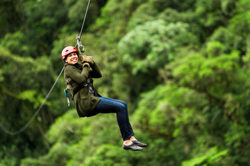 A woman in a helmet and harness ziplines through the dense green jungle canopy in Ecuador, suspended mid-air with a smile, capturing the thrill of an extreme canopy adventure. © Ammit