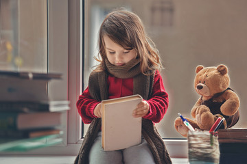 Adorable little girl, writing letter to Santa, sitting on a window, books and teddy bear around