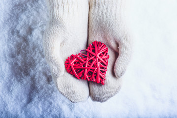 Female hands in white knitted mittens with entwined vintage romantic red heart on snow background....