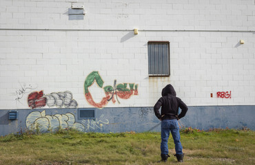 horizontal image of a man in black hoodie standing and looking at graffiti painted on an old exterior wall.