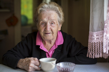 Old woman drinking tea sitting in the kitchen.