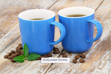 Welcome home card with two mugs of coffee on rustic wooden surface
