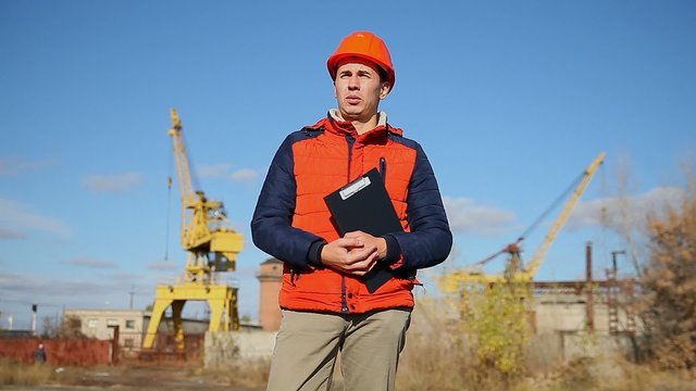 man construction worker in an orange helmet holding documents in background blue sky and crane
