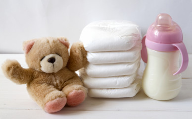 New born child stack of diapers, tebby bear toy and baby feeding bottle with milk on a white wooden...