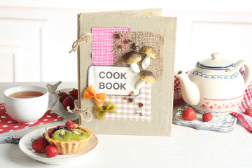 Decorated cookbook with teapot and tasty cake on white background