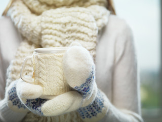 Woman hands in white and blue mittens holding a cozy knitted cup with hot cocoa, tea or coffee. Winter and Christmas time concept.