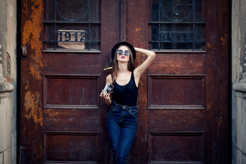 young beautiful brunette girl in a hat, sunglasses, t-shirt and jeans, with a film camera walks in the old city, holding hand hat for fear of losing it