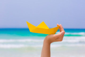 Female hand playing with yellow paper boat in water on the white sand beach on blue sea background