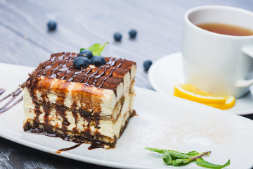 A tiramisu and a cup of tea on the white plate with blueberries and a mint on the woden table