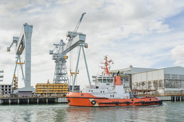 Tugboat at work at a shipyard with cranes in the background
