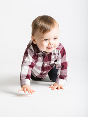 a cute 1 year old baby starts to crawl toward camera in white studio with jeans and a red white flannel looking right