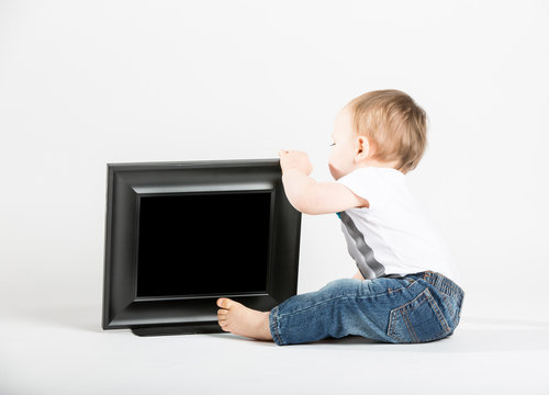 a cute 1 year old baby sits next to a blank black picture frame. He is grabbing the frame and looking behind the picture frame. dressed in Tshirt, jeans, suspenders and blue bow tie