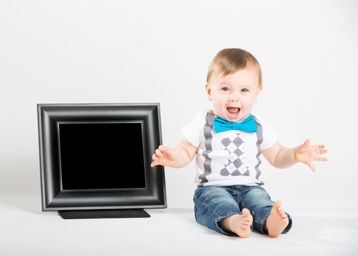 a cute 1 year old baby sits next to a blank black picture frame in a white studio setting. The boy is extremely excited and screaming. He is dressed in Tshirt, jeans, suspenders and blue bow tie