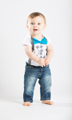 a cute 1 year old stands in a white studio setting. The boy has a interested expression. He is dressed in Tshirt, jeans, suspenders and blue bow tie