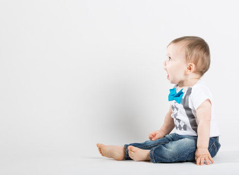 a cute 1 year old stands in a white studio setting. The boy is yelling with an open mouth. He is dressed in Tshirt, jeans, suspenders and blue bow tie