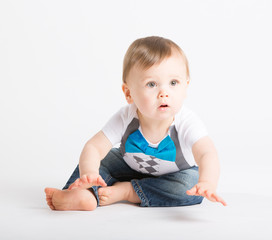 a cute 1 year old sits in a white studio setting. The boy looks like he is about to start crawling. He is dressed in Tshirt, jeans, suspenders and blue bow tie