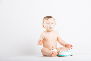 a cute 1 year old sits in a white studio setting. The boy is enjoying his birthday cake with his...