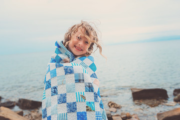 happy child girl at sea wrapped in blanket, warm cozy mood on summer vacation
