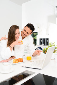 Young Couple In Bathrobe At Home Having Coffee In The Kitchen And Working On Laptop Computer