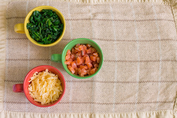 Ingredients for recipe in colorful cups on lien tablecloth. Cubes of smoked salmon in green cup, grated Gouda cheese - in red, cutting spinach - in yellow.  Aerial view