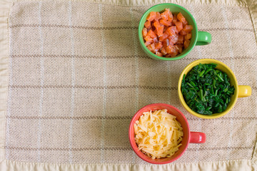 Ingredients in colorful cups on lien tablecloth. Cubes of smoked salmon in green cup, grated Gouda cheese - in red, cutting spinach - in yellow. Look like Traffic Light.  Aerial view